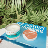 It's Paradise® "Banzai Pipeline" Soy Candle sitting on a surfing book with tropical flora in the background 