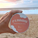 It's Paradise® "Banzai Pipeline" Soy Candle held in a hand with a sandy beach and the ocean in the background